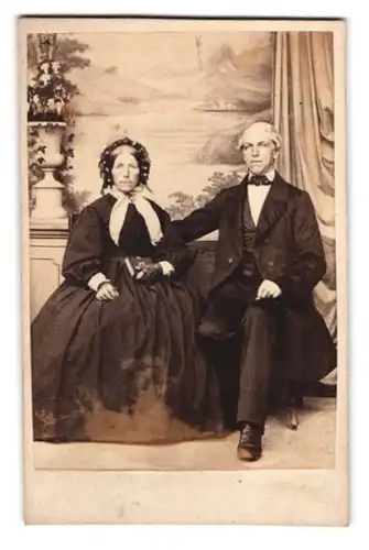 Fotografie unbekannter Fotograf und Ort, älteres paar im Anzug und im dunklen Kleid mit Haube vor einer Studiokulisse