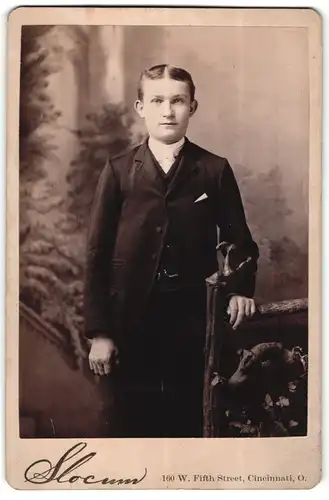 Fotografie Slocum, Cincinnati, O., Portrait elegant gekleideter Knabe an Zaun gelehnt