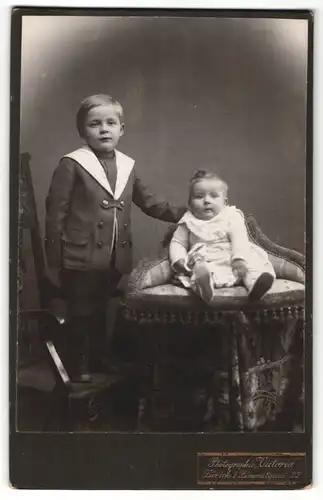Fotografie Victoria, Zürich, Portrait hübsch gekleideter Junge auf Stuhl stehend mit Kleinkind