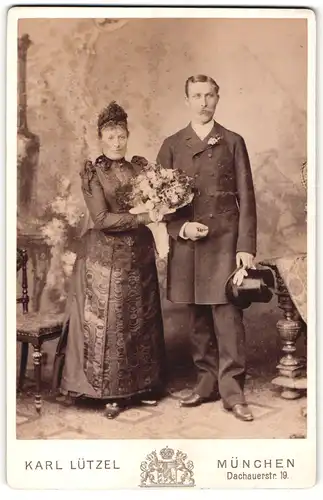 Fotografie Karl Lützel, München, Portrait bürgerliches Paar in hübscher Kleidung mit Zylinder und Blumenstrauss
