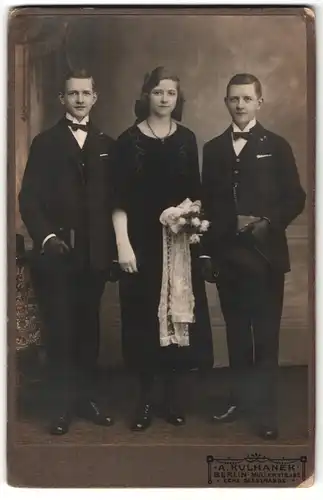 Fotografie A. Kulhanek, Berlin, Portrait junge Dame und zwei Herren in eleganter Kleidung mit Blumen und Gebetsbüchern