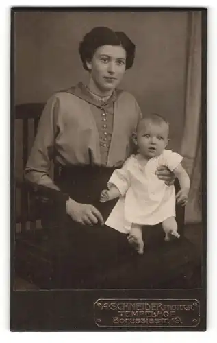 Fotografie A. Schneider, Berlin-Tempelhof, Portrait elegant gekleidete Dame mit Baby auf dem Schoss