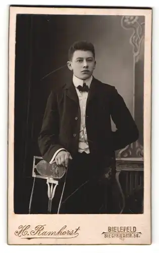 Fotografie H. Ramhorst, Bielefeld, Portrait junger Mann im Anzug mit Fliege an Stuhl gelehnt