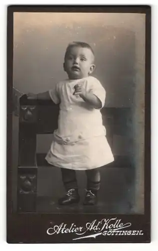 Fotografie Ad. Kolle, Göttingen, Portrait niedliches Kleinkind im weissen Kleid auf Bank stehend