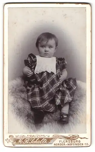 Fotografie M. B. Schultz, Flensburg, Portrait niedliches Kleinkind im karierten Kleid auf Fell sitzend
