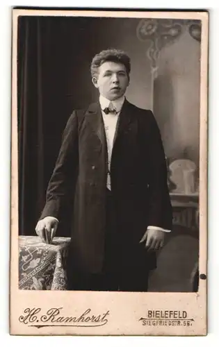 Fotografie H. Ramhorst, Bielefeld, Portrait junger Mann in eleganter Kleidung mit Büchlein an Tisch gelehnt