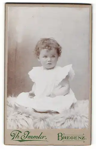 Fotografie Th. Immler, Bregenz, Portrait niedliches Kleinkind im weissen Kleid auf Fell sitzend
