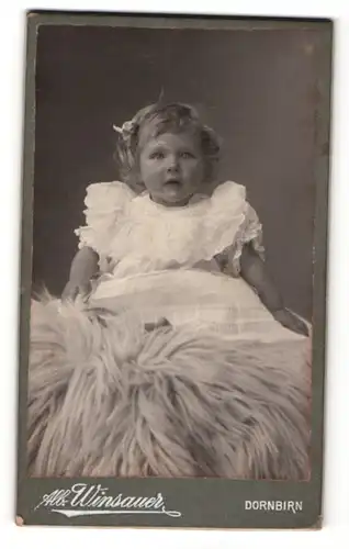 Fotografie Alb. Winsauer, Dornbirn, Portrait niedliches Kleinkind im weissen Kleid auf Fell sitzend