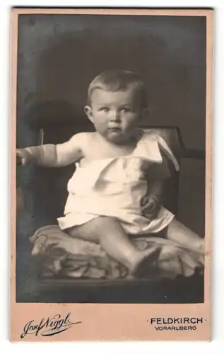 Fotografie Josef Niggl, Feldkirch / Vorarlberg, Portrait niedliches Kleinkind im weissen Hemd auf Kissen sitzend
