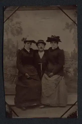 Fotografie Ferrotypie junger Mann mit zwei Damen auf dem Schoss vor einer Studiokulisse