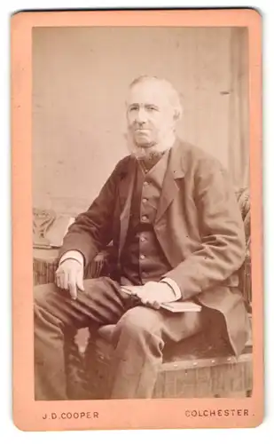 Fotografie J. D. Cooper, Colchester, Portrait bürgerlicher Herr mit weissem Bart und Buch am Tisch sitzend