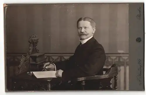 Fotografie A. Jandorf & Co., Berlin, Herr mit Schnauzbart und dunklem Anzug sitzt mit einem Stift in der Hand an Tisch