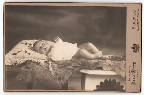 Fotografie Otto Witte, Berlin, Baby in weissem Laibchen liegt auf einem Kissen und einem Fell