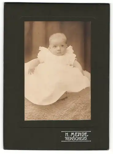 Fotografie H. Mende, Remscheid, Portrait niedliches Baby im weissen Kleid auf Decke sitzend