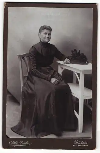 Fotografie Wilh. Leube, Geithain, Portrait elegant gekleidete Dame mit Hut am Tisch sitzend