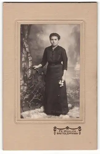 Fotografie Fr. Mühlich, Bretten, Dame in schwarzem Kleid hält weisse Blumen und steht auf einem Fellteppich