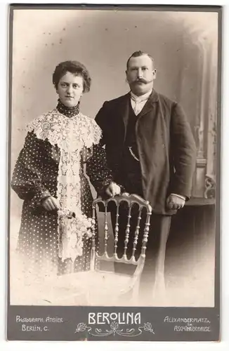 Fotografie Berolina, Berlin-C, Portrait modisch gekleidetes Paar mit Blumen an Stuhl glehnt