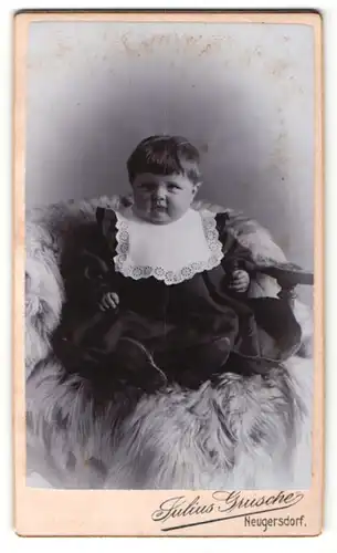 Fotografie Julius Grusche, Neugersdorf i. / Sachs., Portrait hübsch gekleidetes Kleinkind mit Latz auf Fell sitzend
