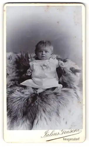 Fotografie Julius Grusche, Neugersdorf i / Sachs., Portrait süsses Kleinkind im weissen Kleid auf Fell sitzend