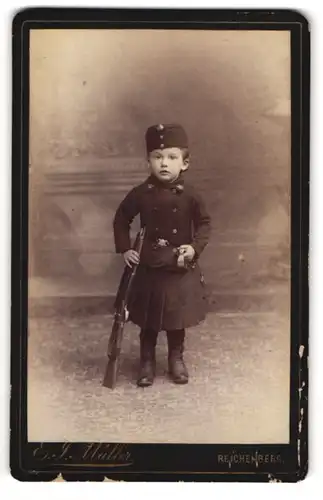 Fotografie Ernst J. Müller, Reichenberg / Liberec, niedliches kleines Mädchen als Soldatin in Uniform mit Gewehr bei Fuss