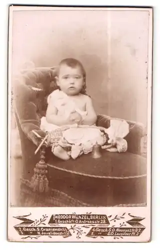 Fotografie Theodor Wenzel, Berlin, Portrait sitzendes Kleinkind im weissen Hemd mit nackigen Füssen und Glocke