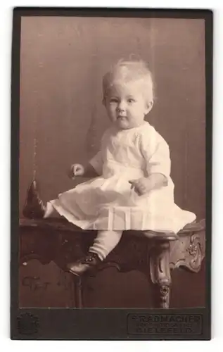 Fotografie F. Radmacher, Bielefeld, Portrait süsses Kleinkind im weissen Kleid auf Tisch sitzend