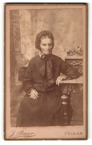 Fotografie J. Baur, Colmar, Ältere Dame mit dunkler Haube sitzt an einem Tisch