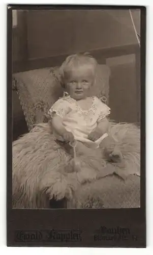 Fotografie Ewald Kappler, Bautzen, Portrait süsses Kleinkind im weissen Hemd mit Rassel auf Fell sitzend