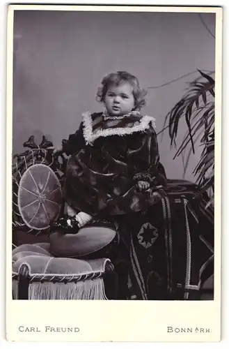 Fotografie Carl Freund, Bonn a / Rh., Portrait niedliches Kleinkind im Samtkleid auf Tisch sitzend