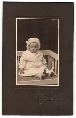 Fotografie Carl Huth, Lüdenscheid, Portrait sitzendes Kleinkind in hübscher Kleidung mit Mützchen
