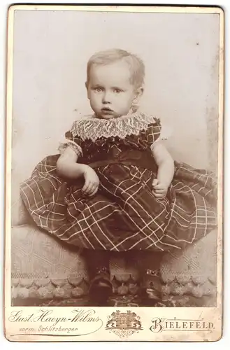 Fotografie Gust. Haeyn-Wilms, Bielefeld, Portrait sitzendes Kleinkind im karierten Kleid mit Spitzenkragen