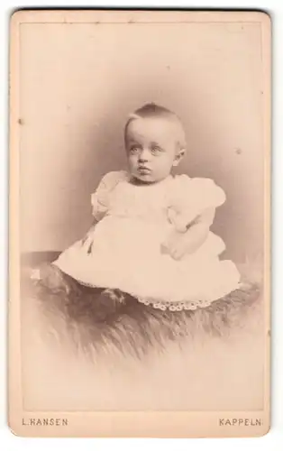 Fotografie L. Hansen, Kappeln, Portrait blondes süsses Kleinkind im weissen Kleidchen auf Fell sitzend