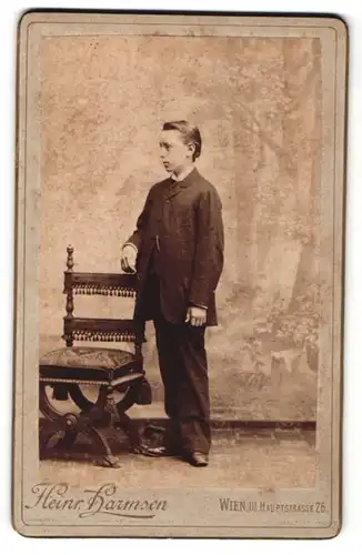 Fotografie Heinr. Harmsen, Wien, Portrait niedlicher Bube im Anzug am Stuhl stehend
