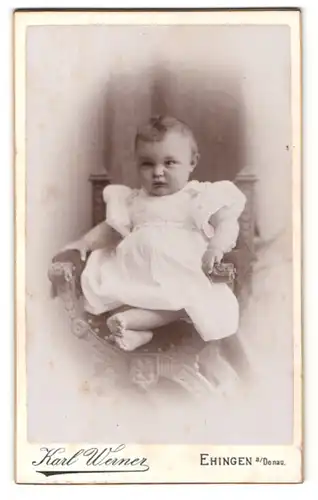 Fotografie Karl Werner, Ehingen a / Donau, Portrait sitzendes Kleinkind im weissen Kleid mit nackigen Füssen