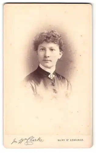 Fotografie W. Clarke, Bury St. Edmunds, Portrait schöne Frau mit lockigem Haar und Brosche am Blusenkragen