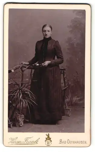 Fotografie Herm. Frank, Bad Oeynhausen, Portrait elegant gekleidete Frau mit Blume am Holzgeländer stehend