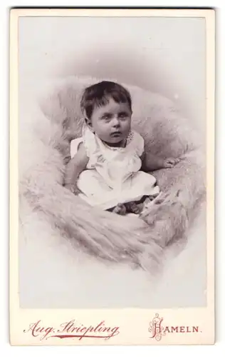 Fotografie Aug. Striepling, Hameln, Portrait dunkelhaariges kleines Mädchen im weissen Hemdchen auf Fell sitzend