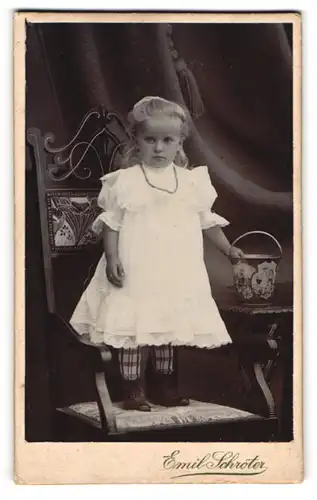Fotografie Emil Schröter, Jüterbog, niedliches Mädchen im weissen Kleid mit Perlenkette steht auf einem Stuhl
