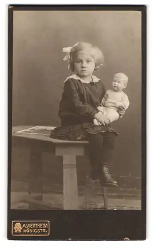 Fotografie A. Wertheim, Berlin, niedliches Mädchen im Kleid mit ihrer Puppe im Arm, Haarschleife