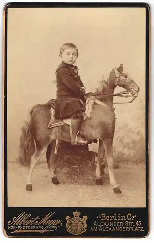 Fotografie Albert Meyer, Berlin, junger Knabe im Samtkleid auf grossen Spielzeug Pferd im Atelier