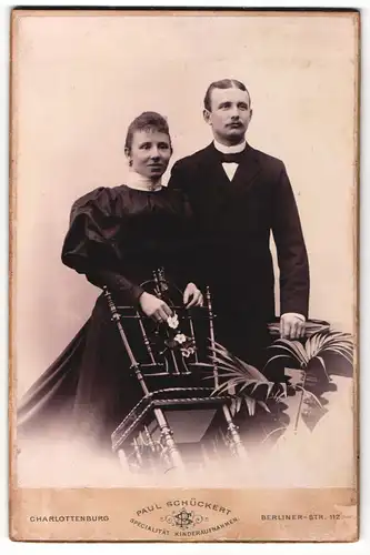 Fotografie Paul Schückert, Berlin-Charlottenburg, Portrait elegant gekleidetes Paar mit Blumen an Stuhl gelehnt
