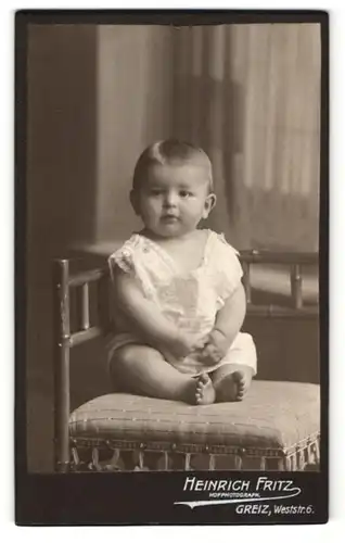 Fotografie Heinrich Fritz, Greiz, Portrait sitzendes Kleinkind im weissen Hemd mit nackigen Füssen