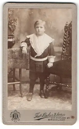 Fotografie Thos Baker & Son, Birmingham, Portrait kleiner Junge in hübscher Kleidung an Tisch gelehnt