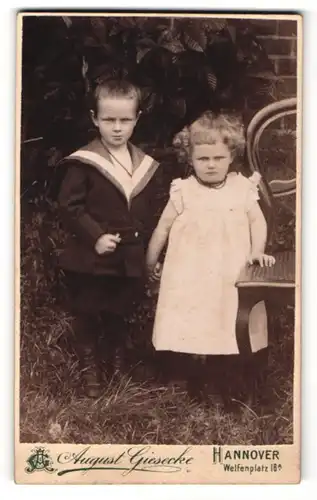 Fotografie August Giesecke, Hannover, Portrait Kinderpaar in hübscher Kleidung an Stuhl gelehnt