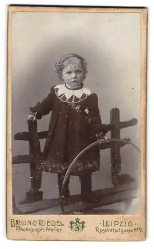 Fotografie Bruo Riedel, Leipzig, Rosenthalgasse 9, Niedliches Mädchen im dunklen Kleid stehend auf Bank mit Reifen