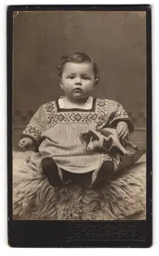 Fotografie F. Lerchenmüller, Kempten, Niedliches Baby in gestreiften Kleid mit Spielzeugkuh und erwartungsvollem Blick