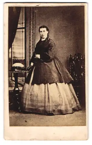 Fotografie unbekannter Fotograf und Ort, junge Dame im hellen Kleid mit Überwurf und Federhut auf dem Tisch