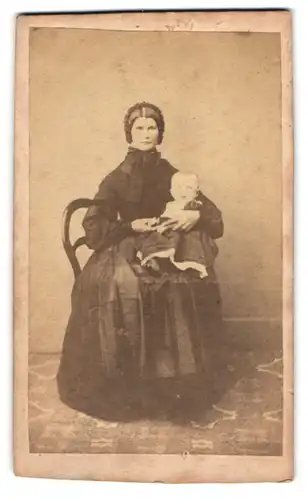 Fotografie unbekannter Fotograf und Ort, streng schauende Mutter mit ihrem Kind auf dem Schoss