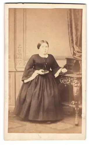 Fotografie unbekannter Fotograf und Ort, Kleinwüchsige Dame im schwarzen Kleid mit Spitze stehend am Sekretär