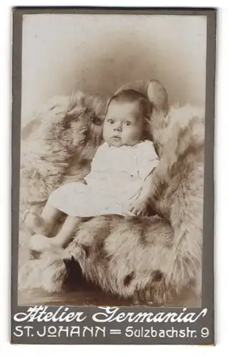 Fotografie Atelier Germania, St. Johann, Portrait süsses Baby im weissen Kleid auf Fell sitzend